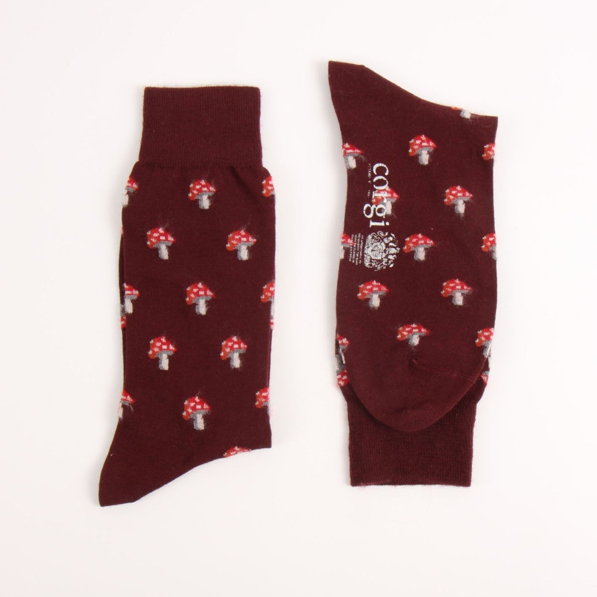A pair of dark burgundy crew socks with a repeating red and white mushroom print, shown flat on a white background. One sock is oriented vertically to show the side pattern, and the other is horizontal showing the toe and the brand name 'corgi' printed on the side.