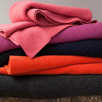 Stack of colourful throws including pink, purple, red, and black on a wooden surface.