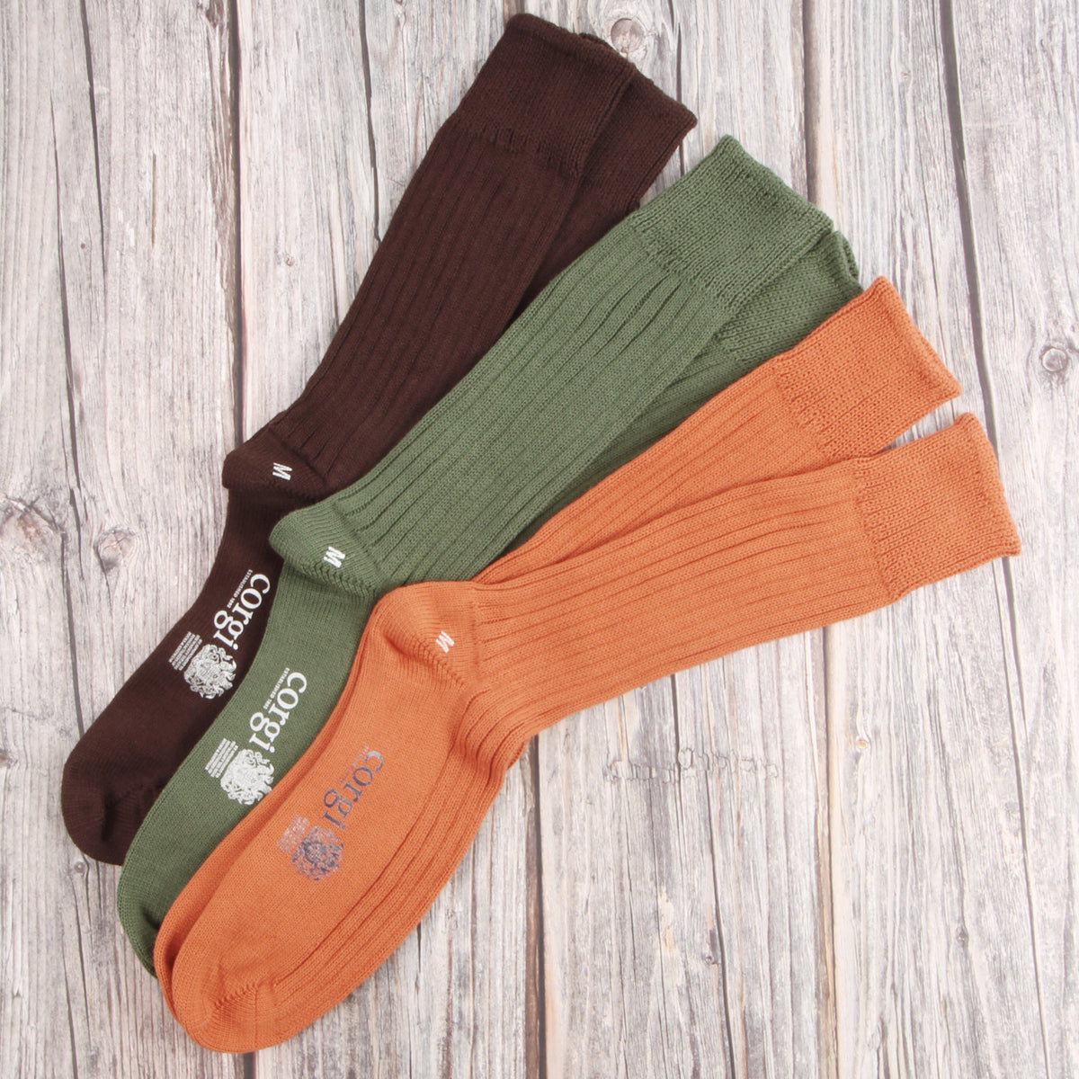 Four ribbed socks in brown, olive green, and orange, arranged diagonally on a light gray wood plank background. The socks feature the brand name 'CORGI' printed near the cuff.