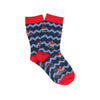 A pair of kids' socks with a snowboarding theme, featuring a zigzag pattern with red and white stripes and a snowboard graphic, placed on a wooden surface with pine branches in the background.